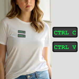Puede incluir: Una camiseta blanca de manga corta con dos parches rectangulares de color gris oscuro en el pecho. El parche superior muestra el texto verde CTRL C y el parche inferior muestra el texto verde CTRL V, imitando comandos de teclado.