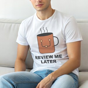 Puede incluir: Camiseta blanca con un gráfico de taza de café marrón y el texto "REVIEW ME LATER" en negro. El diseño de la taza incluye vapor y un diseño similar a una cara. La camiseta es de manga corta y de material suave.
