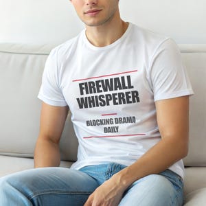 Op de afbeelding: Wit t-shirt met de tekst "FIREWALL WHISPERER" in zwart, met rode lijnen erboven en eronder. Daaronder staat "BLOCKING DRAMA DAILY" in zwart. De persoon draagt een spijkerbroek.