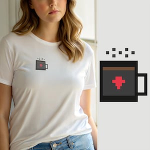Puede incluir: Una camiseta blanca de manga corta con un pequeño gráfico de pixel art en el pecho izquierdo. El gráfico muestra una taza gris oscuro llena de líquido marrón, con una cruz roja pixelada y pequeños cuadrados grises que representan vapor.