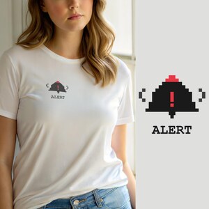 Op de afbeelding: Een wit T-shirt met korte mouwen en een kleine pixel art afbeelding op de borst. De afbeelding toont een zwarte klokvorm met een rood uitroepteken erin, geflankeerd door twee haakjes, met het woord ALERT in het zwart eronder gedrukt.