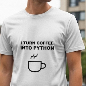Op de afbeelding: Wit T-shirt met de tekst "I TURN COFFEE INTO PYTHON" en een afbeelding van een koffiekopje. Het shirt met korte mouwen is waarschijnlijk van katoen. De achtergrond toont een vage stedelijke omgeving.