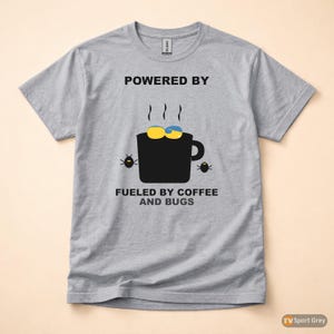 Puede incluir: Camiseta gris con el texto "POWERED BY" sobre un gráfico de taza de café negra con detalles amarillos y azules, vapor y dos ilustraciones de insectos. El texto "FUELED BY COFFEE AND BUGS" está debajo.