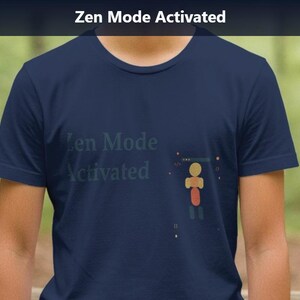 Op de afbeelding: Marineblauw t-shirt met de tekst "Zen Mode Activated" in lichtblauw. Het shirt heeft een kleine afbeelding van een persoon met een rode torso en gele kop. De achtergrond is een vage bosscène.