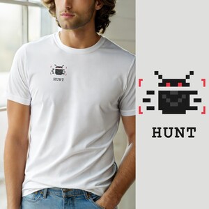 Könnte beinhalten: Ein weißes T-Shirt mit kurzen Ärmeln und einer kleinen, verpixelten Grafik auf der linken Brust. Die Grafik zeigt ein schwarzes, insektenartiges Wesen mit roten Augen, eingerahmt von roten Klammern, mit dem Wort HUNT in schwarzer Schrift.