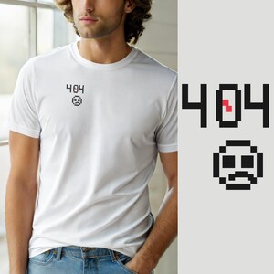 Könnte beinhalten: Ein weißes kurzärmeliges T-Shirt mit einer kleinen schwarzen Pixelgrafik auf der linken Brust. Die Grafik zeigt den Text 404 mit einem roten Pixel im Inneren der Null, platziert über einem pixeligen traurigen Gesicht mit einem Mund.