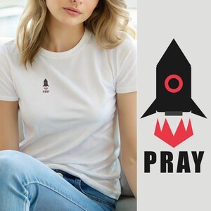 Könnte beinhalten: Ein weißes kurzärmeliges T-Shirt mit einer kleinen Grafik auf der Brust. Die Grafik zeigt eine schwarze Rakete mit einem roten kreisförmigen Fenster und roten Flammen an der Basis, über dem schwarzen Text PRAY in fetter serifenloser Schrift.