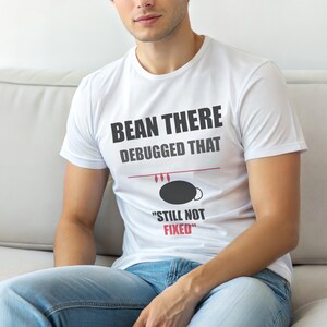 Könnte beinhalten: Weißes T-Shirt mit dem schwarzen Text "BEAN THERE DEBUGGED THAT" und "STILL NOT FIXED" in Rot. Das Design beinhaltet eine Kaffeebohnen-Grafik. Das T-Shirt wird von einer Person getragen.