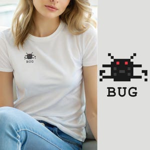 Puede incluir: Una camiseta blanca de manga corta con un pequeño gráfico pixelado en el pecho. El gráfico muestra una criatura negra similar a un insecto con dos ojos rojos brillantes, situada sobre la palabra BUG escrita en una fuente negra, negrita y sin remates.