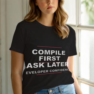 Op de afbeelding: Zwart T-shirt met de witte tekst "COMPILE FIRST ASK LATER", onderstreept met rode lijnen. Daaronder staat de zin "EVELOPER CONFIDENC". Het T-shirt heeft korte mouwen.