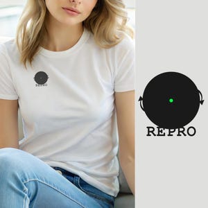 Może przedstawiać: Biały t-shirt z krótkim rękawem z czarną okrągłą grafiką na klatce piersiowej. Grafika przedstawia czarną tarczę z małą zieloną kropką pośrodku, dwiema małymi strzałkami po bokach oraz słowem REPRO wydrukowanym czarnymi wielkimi literami.
