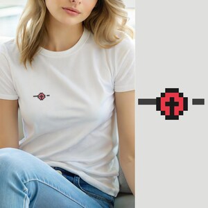 Puede incluir: Una camiseta blanca de manga corta con un pequeño gráfico de pixel art en el pecho izquierdo. El diseño consiste en una forma circular roja con una cruz negra en su interior, flanqueada por dos líneas horizontales negras a los lados.