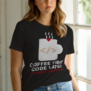 Op de afbeelding: Zwart t-shirt met een witte koffiemok afbeelding, een hart en code symbolen. De tekst luidt "COFFEE FIRST CODE LATER // PRIORITIES COMPILED". Een leuke tee voor programmeurs en koffieliefhebbers.