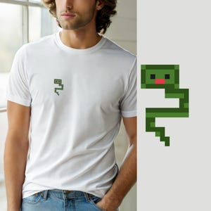 May include: A white short-sleeved t-shirt featuring a small pixel art graphic of a green snake with a red tongue on the left chest area. The snake design is composed of dark and light green square blocks with a distinct blocky, retro aesthetic.