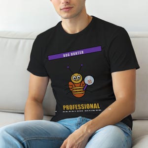 Może przedstawiać: Czarna koszulka z kreskówkową ilustracją robaka trzymającego lupę. Nad robakiem znajduje się napis "BUG HUNTER", a pod nim "PROFESSIONAL PROBLEM FINDER". Projekt jest w kolorze pomarańczowym, fioletowym i białym.