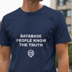 May include: Navy blue t-shirt with white text that reads "DATABASE PEOPLE KNOW THE TRUTH" and a graphic below. The shirt is short-sleeved and appears to be made of a soft material. The background shows a city street.