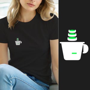 Könnte beinhalten: Ein schwarzes T-Shirt mit kurzen Ärmeln und einer kleinen Grafik auf der linken Brust. Die Grafik zeigt eine weiße Tasse mit Henkel, die drei gestapelte grüne kreisförmige Elemente enthält, sowie ein kleines grünes Rechteck an der Seite.