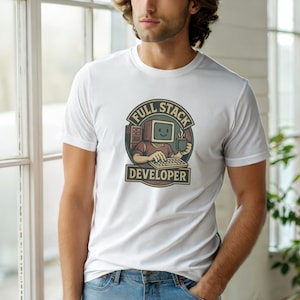 Op de afbeelding: Een wit T-shirt met korte mouwen en een grafische print van een personage met een computerhoofd dat op een toetsenbord typt terwijl het een mok vasthoudt. Het ronde ontwerp bevat de tekst FULL STACK DEVELOPER in een retro kleurenpalet.