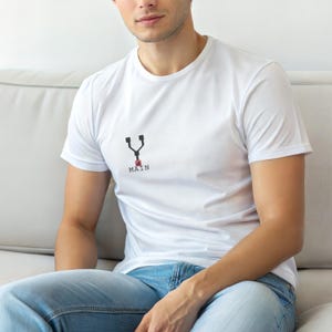 May include: White crew neck t-shirt featuring a black and red graphic and the word "MAIN". The t-shirt is made of a soft material and is worn with blue jeans.