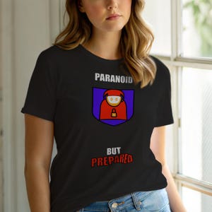 Op de afbeelding: Zwart T-shirt met het woord "PARANOID" boven een schildvormige afbeelding van een persoon in een rode jas en een hangslot. De woorden "BUT PREPARED" staan eronder. Het shirt is gemaakt van een zacht materiaal.