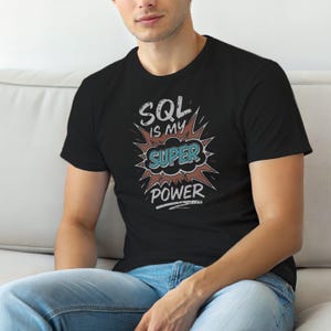May include: A black short-sleeved t-shirt featuring a graphic design with the text "SQL IS MY SUPER POWER" in a comic book style. The word "SUPER" is highlighted in a teal cloud shape, surrounded by a brown starburst and white lettering.