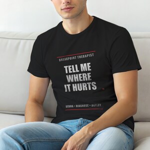 Może przedstawiać: Czarny t-shirt z napisem "BREAKPOINT THERAPIST TELL ME WHERE IT HURTS" w kolorze białym, z napisem "DEBUG • DIAGNOSE • DEPLOY" poniżej. Koszulka jest noszona przez osobę siedzącą na białej kanapie.