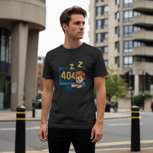May include: Black t-shirt featuring a cartoon illustration of a person sleeping in front of a computer screen displaying "404" error message. The design includes "Z" symbols, suggesting sleep. The shirt is worn by a person.