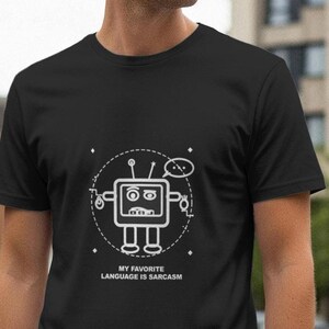 以下が含まれることがあります： 白いロボットのグラフィックと「MY FAVORITE LANGUAGE IS SARCASM」のテキストがプリントされた黒いTシャツ。ロボットは四角い頭、アンテナ、吹き出しが付いています。Tシャツは半袖です。