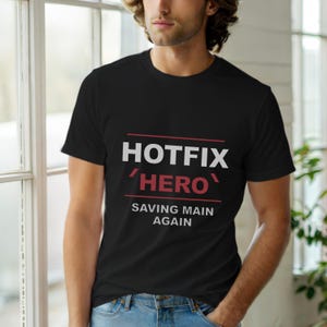 Puede incluir: Camiseta negra con las palabras "HOTFIX HERO" en blanco y rojo, con la frase "SAVING MAIN AGAIN" debajo. La camiseta es de cuello redondo.