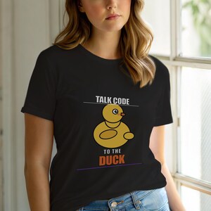 Puede incluir: Camiseta negra con un pato amarillo de dibujos animados y el texto "TALK CODE TO THE DUCK". El pato tiene un cuerpo redondo, un pico pequeño y un solo ojo. El texto es blanco y naranja.