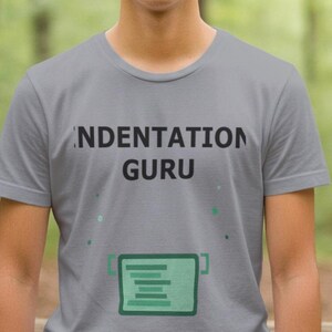 Op de afbeelding: Een grijs T-shirt met ronde hals met de woorden "INDENTATION GURU" in het zwart gedrukt. Onder de tekst staat een groene afbeelding van een codevenster. Het T-shirt wordt gedragen voor een vage bosachtergrond.
