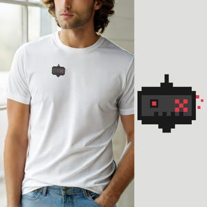 Peut inclure: Un t-shirt blanc à manches courtes avec un petit motif en pixel art sur la poitrine gauche. Le motif représente un personnage rectangulaire gris foncé avec un œil carré rouge et un X rouge pixélisé, accompagné de petits débris rouges.
