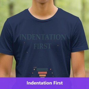 May include: Navy blue t-shirt with the text "INDENTATION FIRST" in a light blue font. The shirt has a crew neck and short sleeves. The bottom of the shirt features a graphic design with orange, red, and blue elements. The background is a blurred forest.