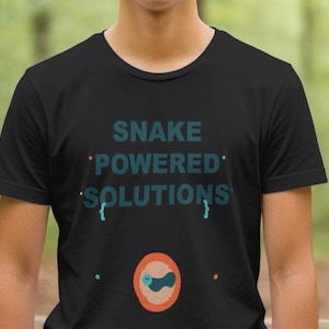 Op de afbeelding: Zwart t-shirt met de tekst "SNAKE POWERED SOLUTIONS" in turquoise. Daaronder een cirkelvormig ontwerp met een slangenillustratie. De achtergrond is een vage bosscène.