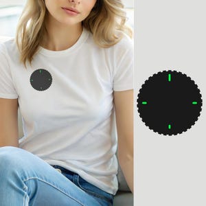 May include: A white short-sleeved t-shirt featuring a circular dark gray graphic on the chest. The graphic has a scalloped edge and four small, bright green rectangular marks positioned at the top, bottom, left, and right sides of the circle.