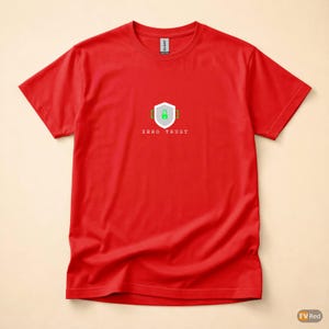 Peut inclure: T-shirt rouge à col rond. Le devant du t-shirt présente un motif blanc et vert d'un bouclier avec un cadenas et le texte "ZERO TRUST". Le t-shirt est en tissu doux.