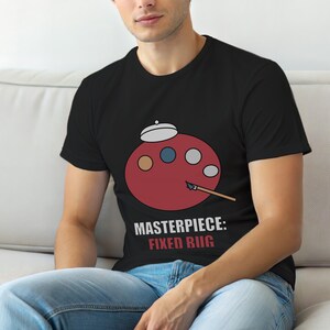 Puede incluir: Camiseta negra con un diseño de paleta de artista roja, con puntos de colores y un pincel. El texto "MASTERPIECE: FIXED BUG" está impreso debajo. La camiseta está hecha de un material suave.
