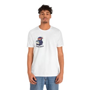 Könnte beinhalten: Weißes T-Shirt mit einem Cartoon-Ninja in schwarzer Kleidung und rotem Stirnband, der an einem Laptop arbeitet. Der Text "CODE NINJA" ist darunter gedruckt. Das Shirt ist aus weichem Material.