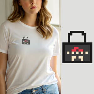 Op de afbeelding: Een wit T-shirt met korte mouwen en een kleine pixel art afbeelding op de borst. De afbeelding is een donkergrijze rechthoekige vorm met een handvat, met een rood en beige gepixeld ontwerp dat lijkt op een paddenstoel of wezen.
