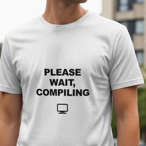 May include: White t-shirt with the text "PLEASE WAIT, COMPILING" in black, along with a small computer monitor icon. The shirt is short-sleeved and appears to be made of a soft material. The shirt is being worn outdoors.