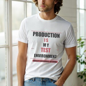 Op de afbeelding: Wit T-shirt met de tekst "PRODUCTION IS MY TEST ENVIRONMENT" in zwart en rood. De zin "QUALITY ASSURANCE OPTIONAL" staat onder de hoofdtekst. Het T-shirt wordt door een persoon gedragen.