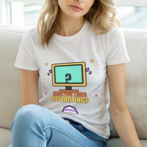 Op de afbeelding: Wit t-shirt met een computermonitorafbeelding en een vraagteken op het scherm. De tekst "HAVE YOU TRIED RESTARTING?" staat eronder. Het shirt heeft ook ster- en stipafbeeldingen.