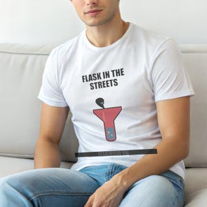 以下が含まれることがあります： 「FLASK IN THE STREETS」の文字と、赤いフラスコのグラフィックがプリントされた白いTシャツ。クルーネックで半袖です。