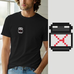 Może przedstawiać: Czarny t-shirt z krótkim rękawem z małą grafiką w stylu pixel art na lewej piersi. Grafika przedstawia ciemnoszary pojemnik z jasnoszarym środkiem i jaskrawoczerwonym, pikselowym kształtem litery X umieszczonym w centrum szarego obszaru.