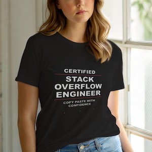 Op de afbeelding: Een zwart t-shirt met korte mouwen en witte tekst met de tekst CERTIFIED STACK OVERFLOW ENGINEER en COPY PASTE WITH CONFIDENCE, geaccentueerd door dunne rode horizontale lijnen en drie kleine rode stippen onder de tekst.