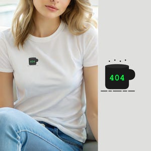 Op de afbeelding: Een wit t-shirt met korte mouwen en een kleine afbeelding op de linkerborst. De afbeelding toont een zwarte koffiemok met de groene tekst 404 op de voorzijde, vergezeld van kleine zwarte stippen boven de mok en een stippellijn eronder.