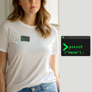 Puede incluir: Una camiseta blanca de manga corta con un pequeño gráfico rectangular en el pecho. El gráfico es una ventana de terminal de computadora gris oscuro con un símbolo del sistema verde que muestra el texto printf("here"); en fuente monoespaciada.