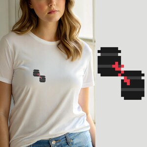 May include: A white short-sleeved t-shirt featuring a small pixel art graphic on the chest. The design consists of two dark gray cylindrical containers connected by a jagged red line, rendered in a simple, blocky digital aesthetic.
