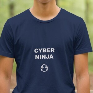 May include: Navy blue t-shirt with the words "CYBER NINJA" in white block letters, along with a white graphic below. The shirt is a crew neck style and appears to be made of a soft material. The background is a blurred forest scene.