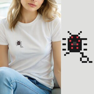 Pode incluir: Uma t-shirt branca de manga curta com um pequeno gráfico de pixel art no peito. O gráfico retrata um inseto preto semelhante a um besouro com pontos vermelhos e uma linha, num estilo digital de blocos simples e limpo.